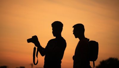 Valokuva kahdesta miehestä, joiden mustat siluetit piirtyvät oranssia taivasta vasten. Toinen miehistä pitelee digitaalikameraa koholla, ja kumpikin miehistä katsoo kameran näyttöä.
