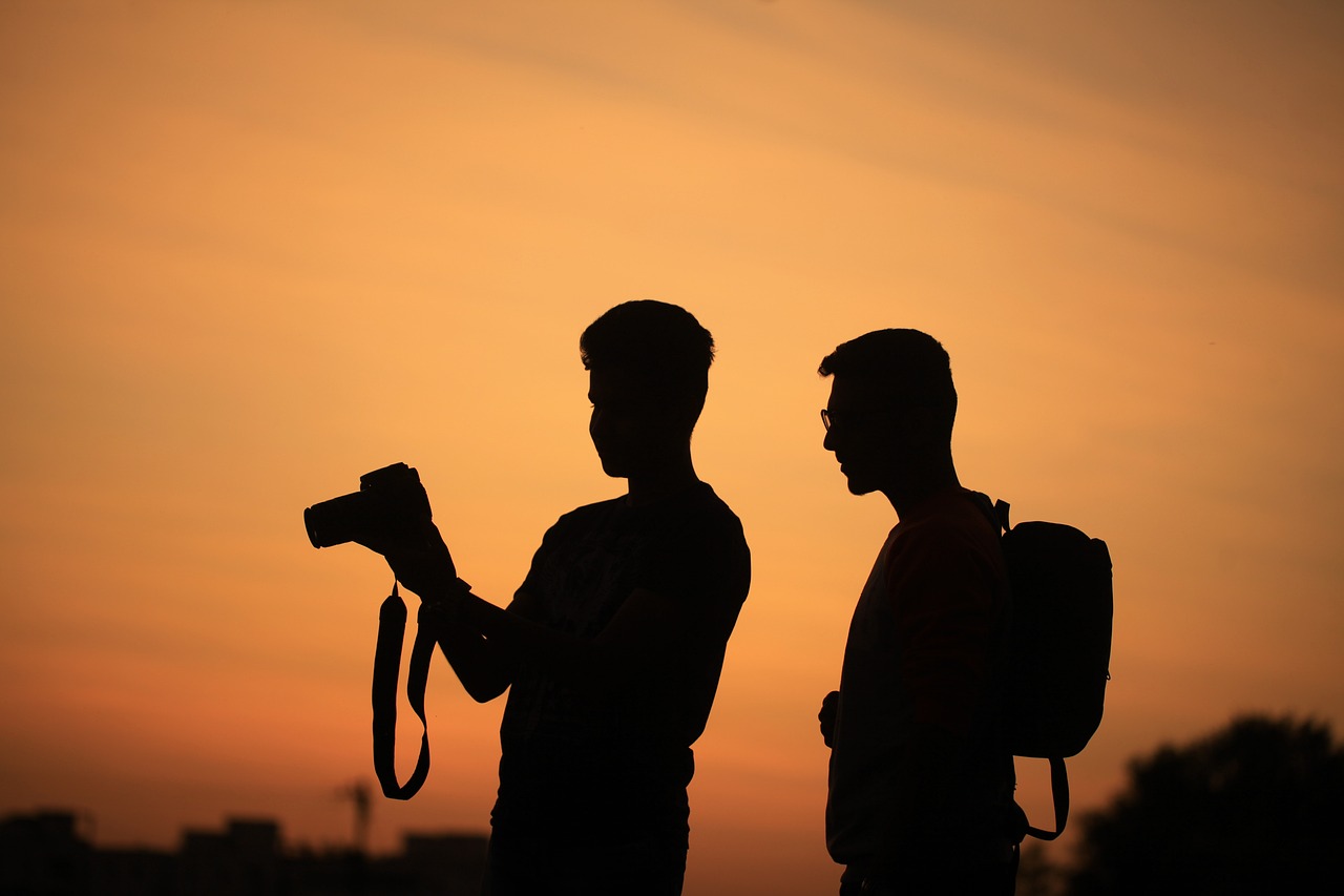 Valokuva kahdesta miehestä, joiden mustat siluetit piirtyvät oranssia taivasta vasten. Toinen miehistä pitelee digitaalikameraa koholla, ja kumpikin miehistä katsoo kameran näyttöä.